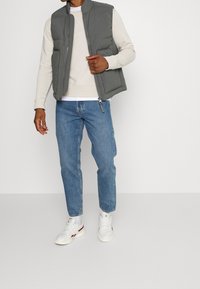 Szary ocieplany kamizelka z wysokim kołnierzem, noszona na beżowym swetrze, wparowana z jasnoniebieskimi dżinsami i białymi trampkami. Widoczne zapięcie na zamek i boczne kieszenie.