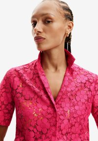 Lys pink blonde skjorte med blomster mønster, korte ærmer og knaplukning foran. Har en spidse krave og en lomme på brystet.