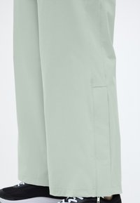 Pantalon ample vert clair avec poche latérale et fermeture éclair près de la cheville, porté avec des chaussures de sport noires et blanches.