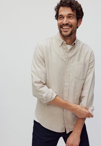 Lichtbeige linnen overhemd met een button-down kraag, lange mouwen opgerold en een borstzakje. Gedragen over een wit shirt en donkere spijkerbroek.