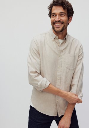 Chemise en lin beige clair avec un col boutonné, manches longues retroussées et une poche poitrine. Portée par-dessus une chemise blanche et un jean foncé.