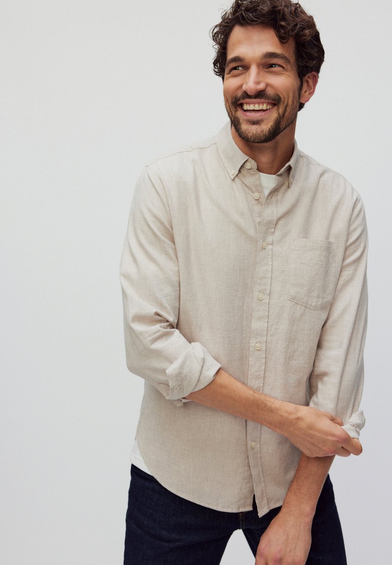 Lichtbeige linnen overhemd met een button-down kraag, lange mouwen opgerold en een borstzakje. Gedragen over een wit shirt en donkere spijkerbroek.