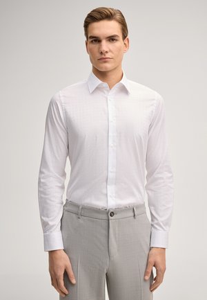 Młody mężczyzna stojący nieruchomo, ubrany w białą koszulę zapinaną na guziki oraz jasnoszare eleganckie spodnie na jednolitym tle.