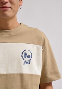 Beige og creme kortærmet t-shirt med et blåt grafisk tryk af en drink og en sandwich, indrammet af et laurbærdesign, på brystet.