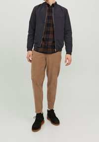 Muž v černé bomber bundě přes hnědou a černou károvanou košili, v béžových kordových kalhotách a černých semišových botách s hnědými podrážkami.