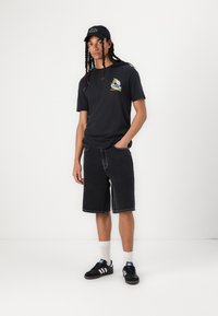 Sort bomulds T-shirt med grafisk tryk, sorte denim shorts og sorte sneakers med hvide striber. Inkluderer en sort kasket med tekst.