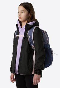 Black jacket with pink accents, lavender hoodie underneath, and a blue backpack with mesh pockets, worn by a girl with medium-length brown hair.