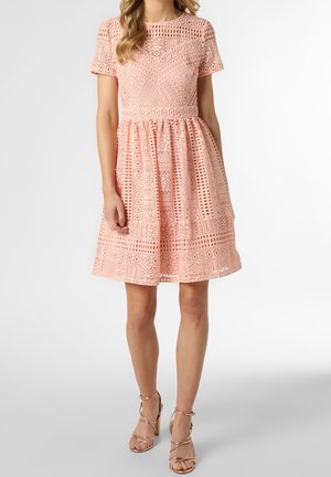 Rozkloszowana sukienka z jasnopomarańczowej koronki o geometrycznych wzorach, z krótkimi rękawami i okrągłym dekoltem, zestawiona z beżowymi sandałkami na paski.