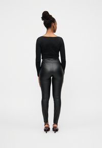 Μαύρο τοπ με μακριά μανίκια και μαύρο κολάν από βινύλιο, εφαρμοστή σχεδίαση, λεία υφή. Άνοιγμα στην πλάτη, φορεμένο με καφέ παπούτσια με τακούνι.