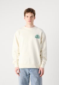 Felpa color crema con un piccolo logo grafico verde sul lato sinistro del petto, collo rotondo, polsini a costine e una vestibilità rilassata. Indossati sotto ci sono jeans in denim.