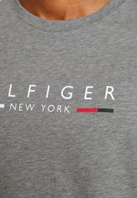 Närbild av grått tyg med vit text "LFIGER NEW YORK" och en liten rektangulär design i vitt, rött och svart.