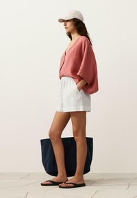 Femeie purtând o bluză coral lejeră, pantaloni scurți albi, șlapi negri și o șapcă bej, stând lângă o geantă tote mare de culoare bleumarin pe podea cu gresie.