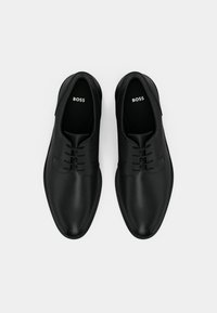 Coppia di scarpe formali nere in pelle con lacci, lati texturizzati e marchio "BOSS" all'interno, su sfondo bianco.