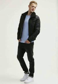 Svart pufferjakke med fuskepelskrage, lys blå skjorte, svarte jeans og hvite sneakers. Jakken har glidelåser og en strukturert passform.