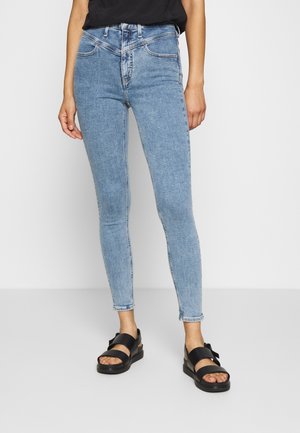 Osoba oblečená v světlých úzkých modrých džínách, černém tílku a černých sandálech s otevřenou špičkou a silnými pásky, stojící na jednobarevném pozadí.