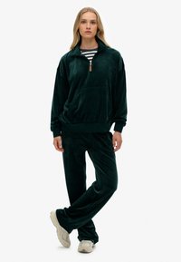 Chándal de terciopelo verde con un pulóver de media cremallera y bolsillo frontal, acompañado de pantalones a juego. Se lleva una camisa de rayas por debajo.