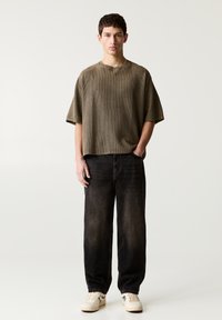 T-shirt oversize a coste verde oliva, vestibilità rilassata; abbinato a jeans ampi neri e sneakers beige con accenti blu.