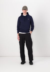 Felpa blu navy con tasca a marsupio, logo giallo sul petto, abbinata a pantaloni cargo neri con tasche laterali, completata con un cappellino grigio.