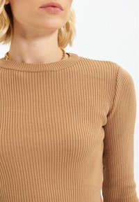 Pull marron côtelé avec un col rond, présentant un tissu texturé. Le design comprend des manches longues et une silhouette cintrée.