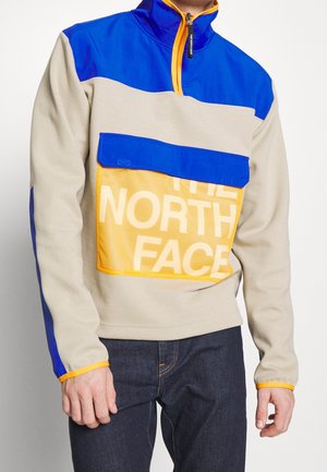 Muž nosící béžovo-modrý pulovr s oranžovými detaily a velkou přední kapsou s nápisem "THE NORTH FACE", v kombinaci s tmavými džínami.