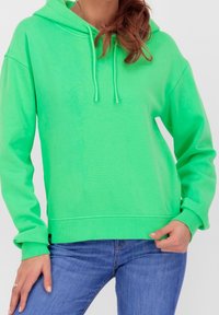 Donna che indossa un felpa verde brillante con cappuccio e jeans blu, mano poggiata vicino alla vita, vista parziale dal mento ai fianchi.