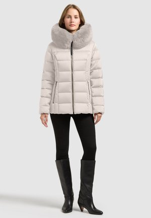 Modèle portant une doudoune blanche matelassée avec un grand col en fourrure grise, un legging noir et des bottes noires montant jusqu’aux genoux.