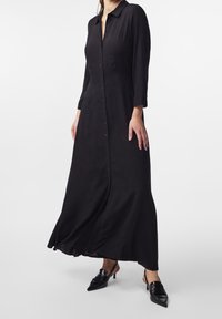 Abito camicia lungo nero con collo a camicia, chiusura con bottoni e gonna fluida. Abbinato a tacchi neri a punta.