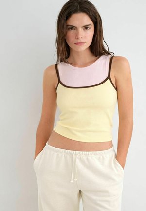 Junge Frau mit braunen Haaren, die ein mehrlagiges ärmelloses Top in Hellrosa und Gelb trägt, kombiniert mit weißen Hosen mit Tunnelzug, stehend mit den Händen in den Taschen.