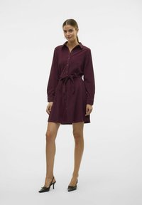 Robe chemise bordeaux avec col, manches longues, taille à nouer et ourlet légèrement évasé. Texture lisse et boutons dorés.
