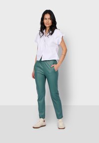 Blusa blanca de manga corta con botones en la parte delantera, combinada con pantalones de cuero sintético verdes que cuentan con bolsillos laterales y un corte ajustado.