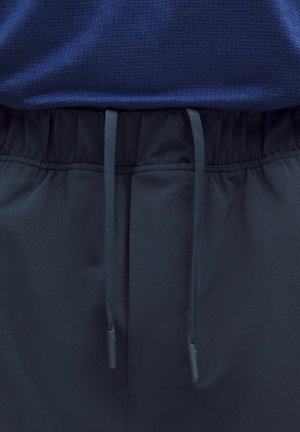 Marineblauwe sportbroek met elastische tailleband en zwarte trekkoorden over een donkerblauw gestructureerd overhemd.