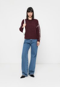 Burgunderstrikkegenser med rynkede detaljer på skuldrene, kombinert med lyseblå vide jeans og svarte flate sko. Hvit veske ligger over skulderen.