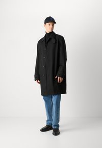 Manteau noir oversize avec de grands boutons, col plat et manches longues ; associé à un jean bleu et des chaussures noires, porté avec une casquette noire.