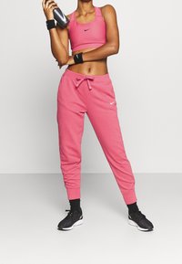 Rosa Nike-Sport-BH und passende Sweatpants, an den Knöcheln gerafft. Schwarze Sportschuhe mit strukturiertem Detail. Wasserflasche in der Hand.