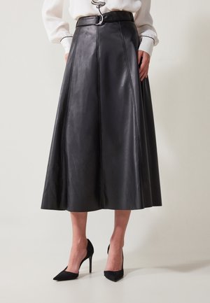 Femme portant une jupe midi en cuir noir trapèze avec ceinture et des chaussures à talons hauts noires à bout pointu, debout devant un fond uni.