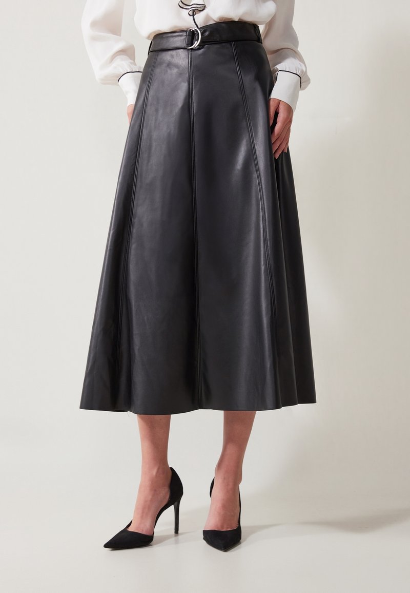 Femme portant une jupe midi en cuir noir trapèze avec ceinture et des chaussures à talons hauts noires à bout pointu, debout devant un fond uni.
