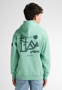 Felpa verde menta con tasca a marsupio frontale e una grande grafica nera con alberi e forme geometriche sul retro.