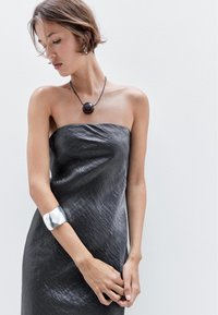 Šedé šaty bez ramínek s metalickým leskem, na jednom zápěstí stříbrný náramek a kolem krku kulatý tmavý přívěsek. Hladká textura.