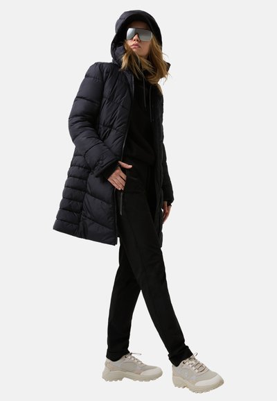 Doudoune noire avec capuche, poches zippées, associée à un sweat à capuche noir et un pantalon noir, complétée par des baskets beiges et des lunettes de soleil surdimensionnées.