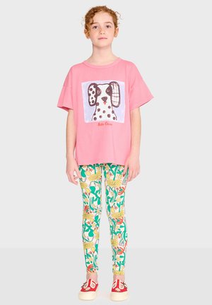 Dievča s kučeravými červenými vlasmi má na sebe ružové tričko s ilustráciou škvrnitého psa, legíny s kvetinovým vzorom a červené nazúvacie topánky.