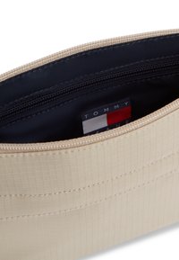 Μπεζ υφασμάτινη θήκη με λείο φερμουάρ. Στο εσωτερικό διαθέτει σκουρόχρωμη υφασμάτινη επένδυση και εμφανή ετικέτα με λογότυπο σε κόκκινες, λευκές και μπλε αποχρώσεις.