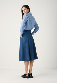 Blau gestreiftes Hemd mit langen Ärmeln, kombiniert mit einem dunkelblauen, ausgestellten Jeansrock. Der Rock hat einen rohen Saum und seitliche Taschen. Schwarze Plateauschuhe.