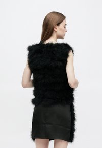 Sort vest i imiteret pels med en tekstureret, lagdelt design; ærmeløs med løs pasform, båret over et glat sort skørt.