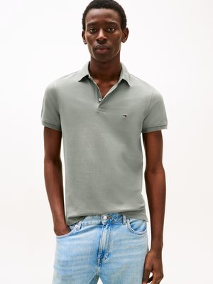 Mann trägt ein hellgraues kurzärmeliges Polohemd mit kleinem Logo und hellblauen Jeans, steht mit einer Hand in der Tasche.