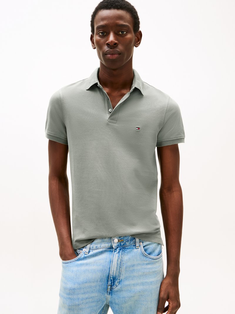 Bărbat purtând un tricou polo gri deschis cu mâneci scurte și un mic logo, și blugi albastru deschis, stând cu o mână în buzunar.