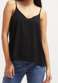 Donna che indossa un top nero senza maniche con scollatura a V e jeans blu in piedi contro uno sfondo bianco semplice.