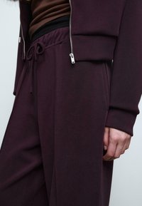 Burgunderfarbene sportliche Zip-Jacke mit Kordelzugbund, ausgestattet mit einer glatten Textur und silbernen Hardware-Akzenten, kombiniert mit dunklen Hosen.