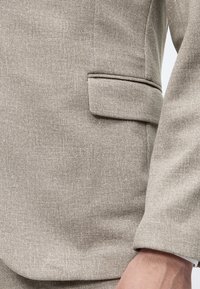 Veste de costume beige texturée avec poche à rabat et manche longue visible sur le bras de la personne.