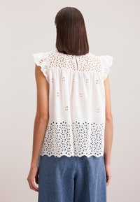 Blusa bianca senza maniche con ricami a occhio, spalline arricciate e orlo scallop. Vestibilità casual, design leggermente morbido.