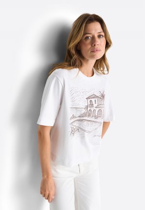 Donna con capelli castano chiaro che indossa una t-shirt bianca con uno schizzo marrone di una casa, una piscina e una palma, abbinata a pantaloni bianchi.